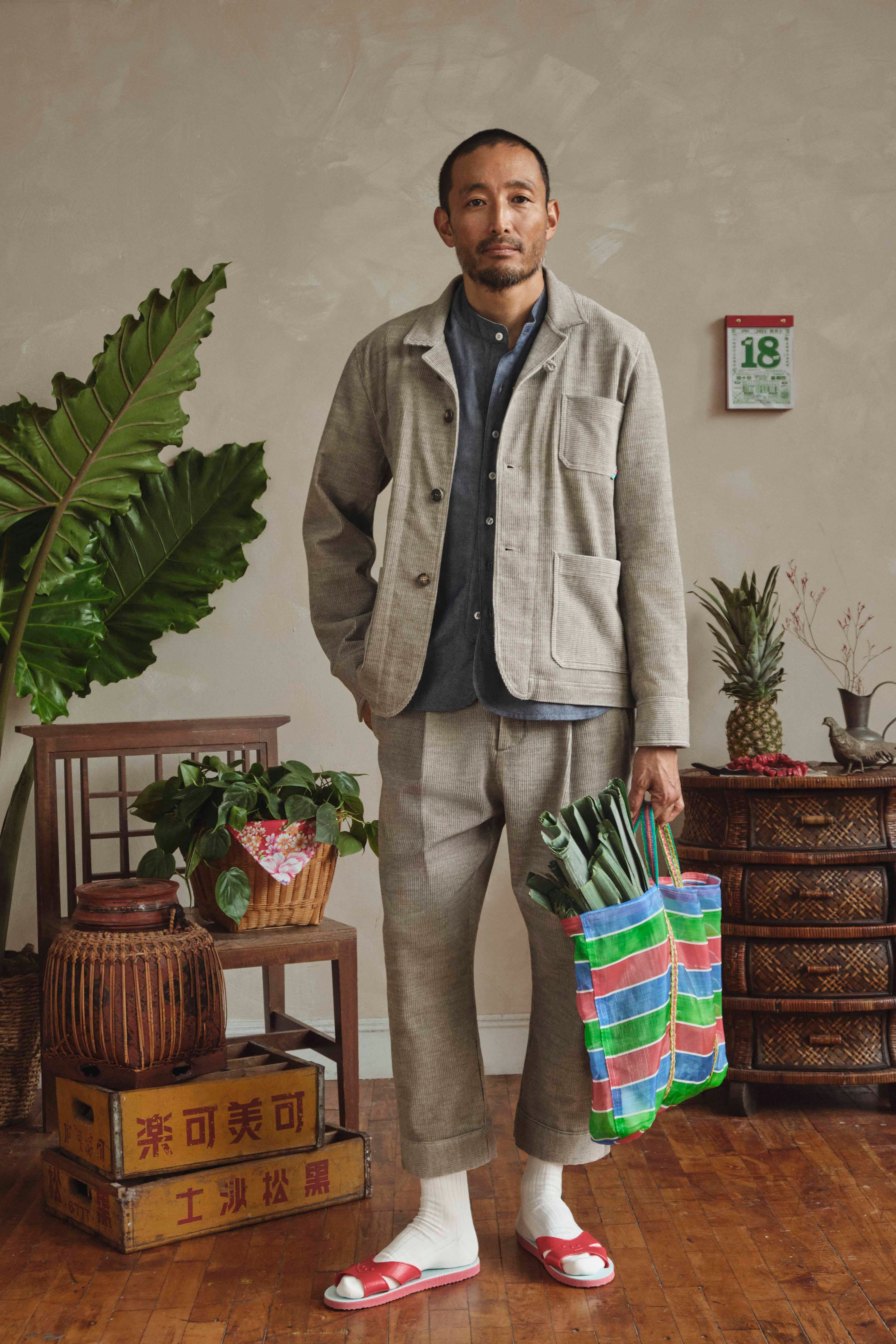 Man wearing a gray corduroy suit holding a colorful bag in a room with plants and furniture