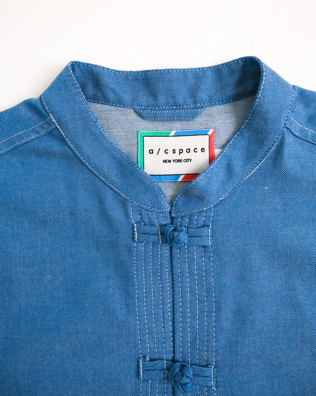 Close-up of a blue denim-like tunic top with a mandarin collar and front button placket, featuring a visible neck label that reads a/cspace NEW YORK CITY.