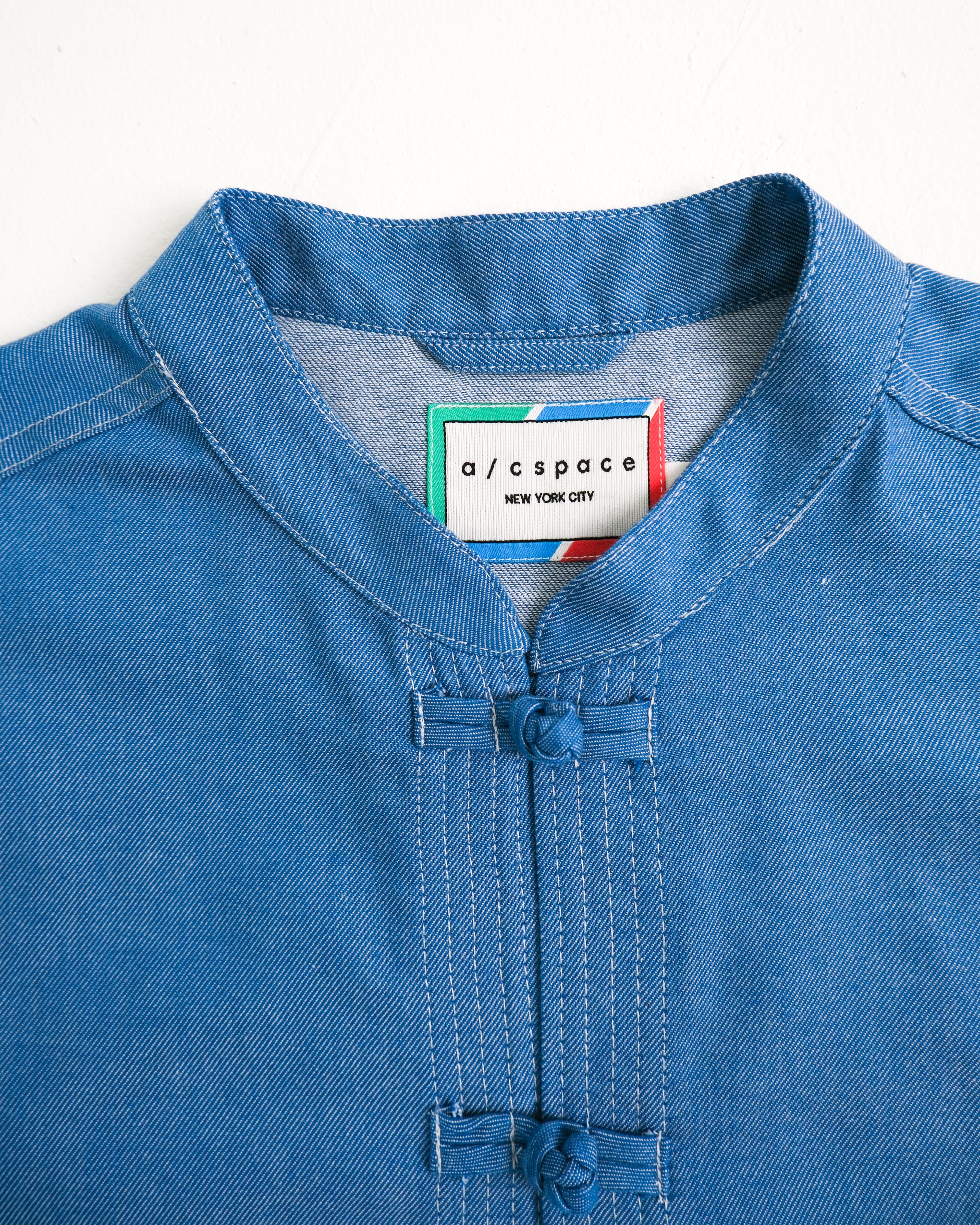 Close-up of a blue denim-like tunic top with a mandarin collar and front button placket, featuring a visible neck label that reads a/cspace NEW YORK CITY.