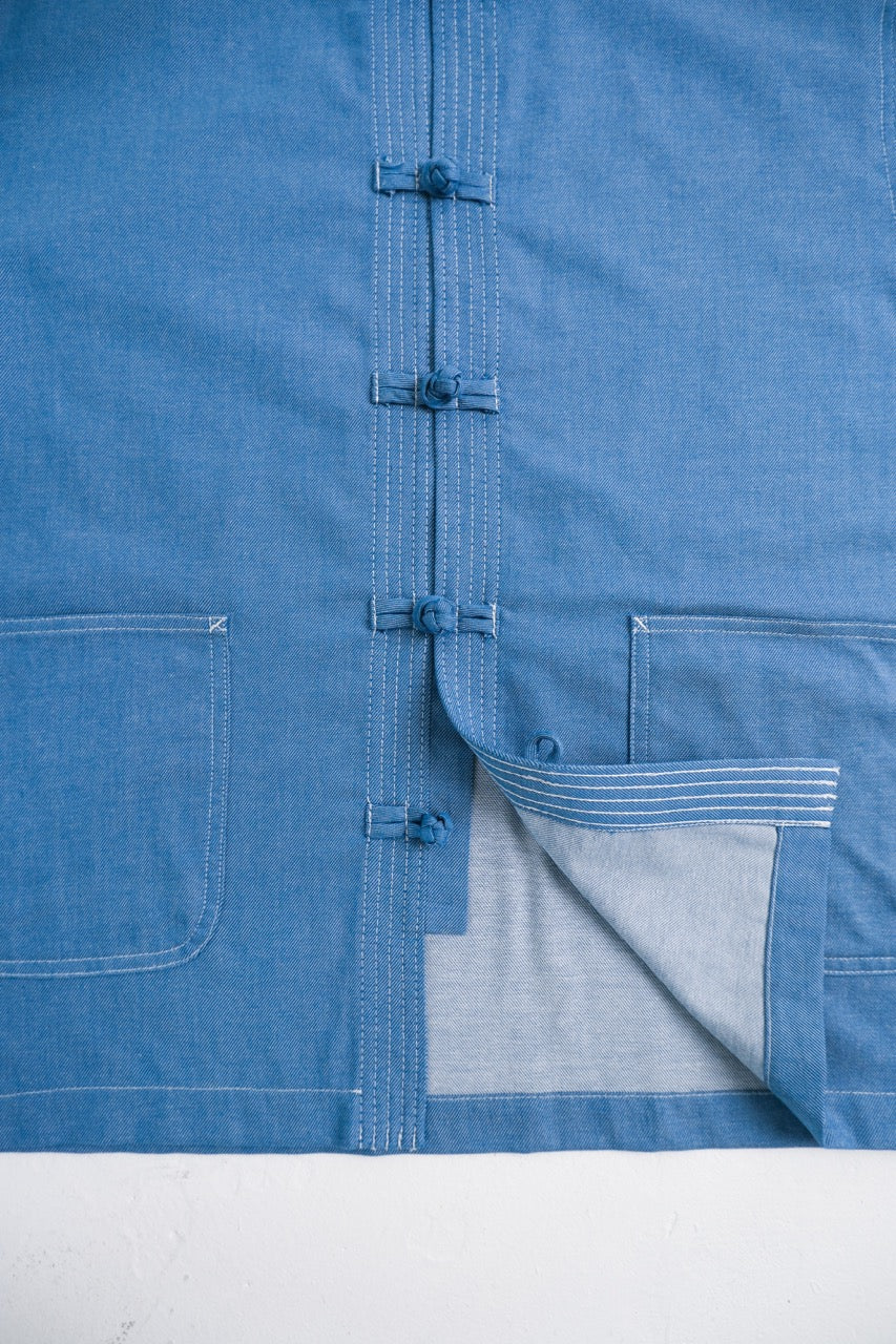 A close-up of a blue fabric garment, likely a shirt or jacket, featuring a central button closure with decorative ties, a pocket on the left, and a pocket on the right with its flap turned down to reveal a lighter lining.