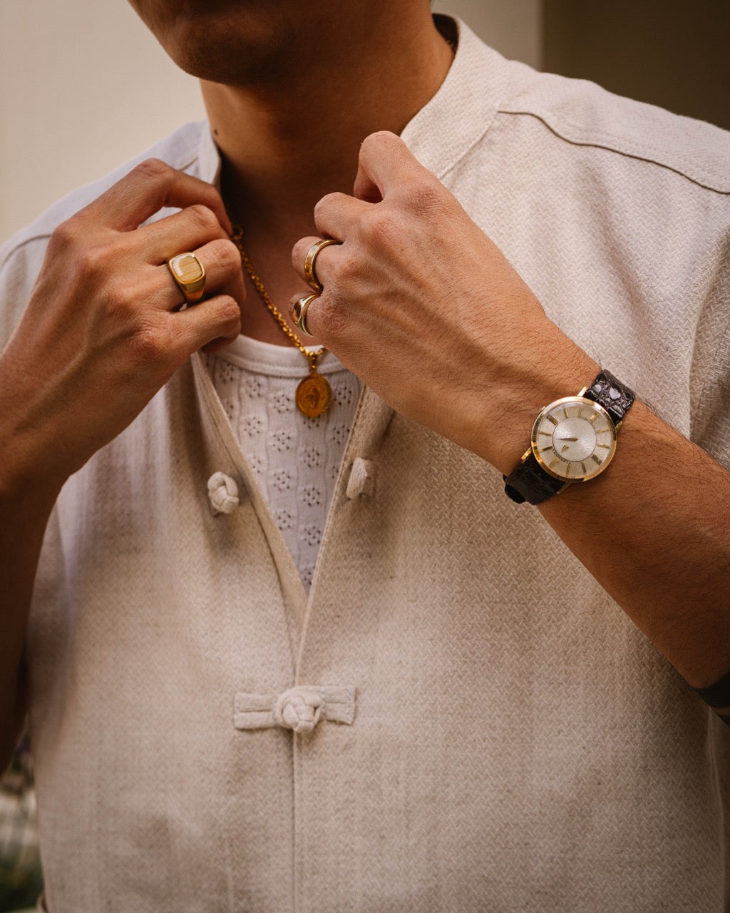 A close-up lifestyle shot of a man wearing a beige linen mandarin collar shirt, adjusting his collar with both hands. He is wearing a gold pendant necklace, gold rings, and a black leather watch with a gold-toned case.