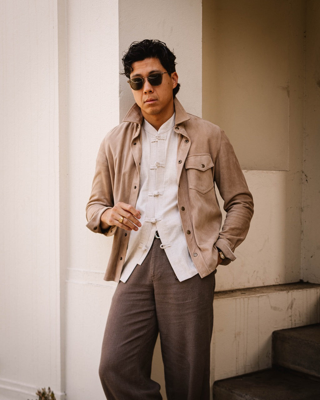 A man wearing a beige suede jacket over a white Chinese-style mandarin collar shirt, paired with brown trousers and sunglasses, standing in front of a white architectural column.