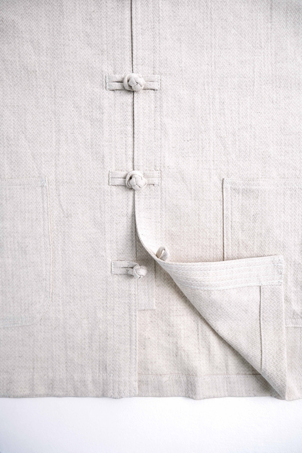 A close-up of a light beige linen garment featuring traditional Chinese-style fabric tie closures and a folded corner showing the textured weave.