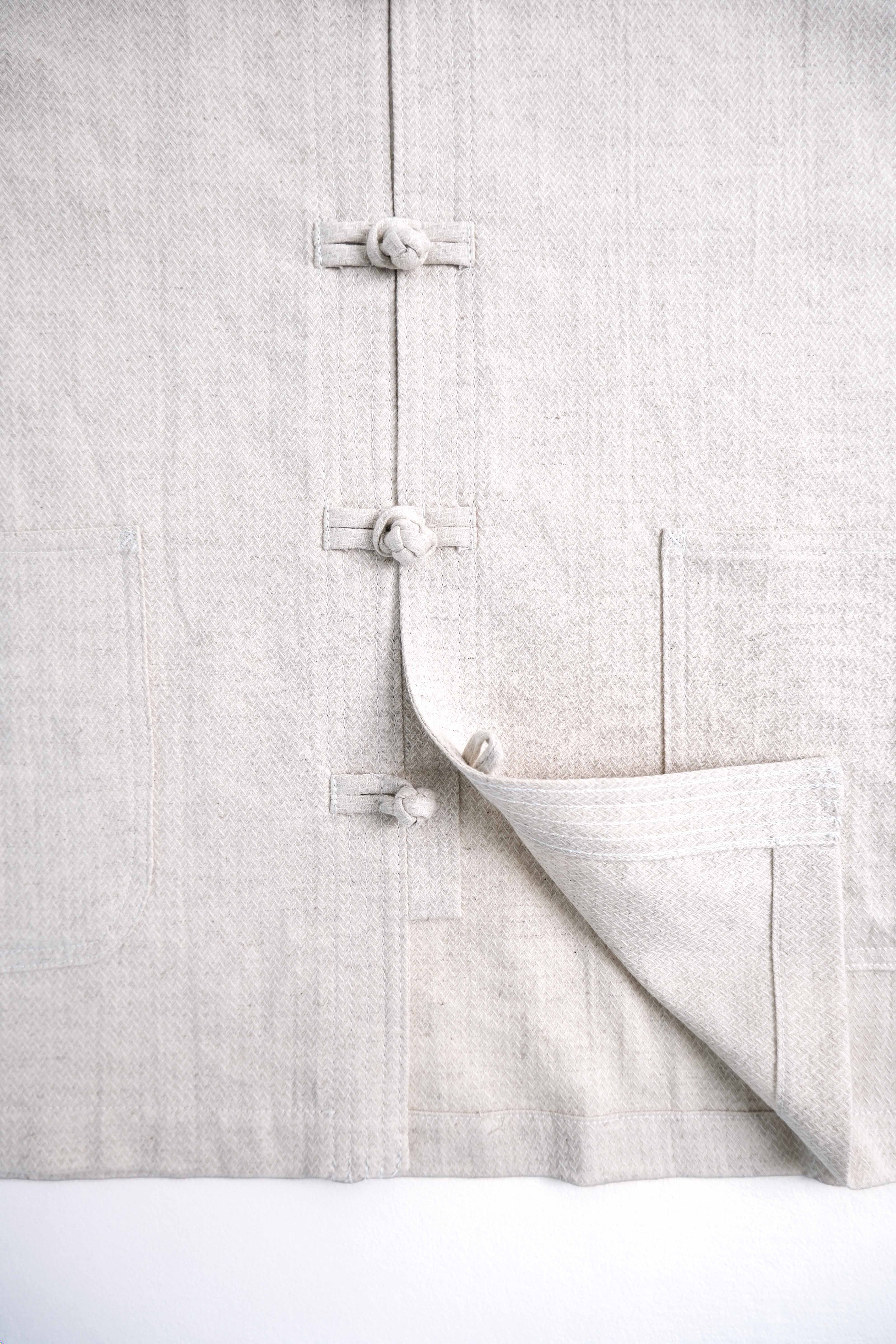 A close-up of a light beige linen garment featuring traditional Chinese-style fabric tie closures and a folded corner showing the textured weave.