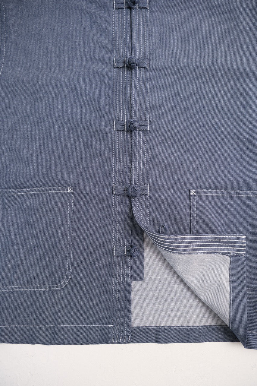 Close-up of a dark indigo blue chambray jacket with traditional Chinese frog closures and a folded hem showing a lighter gray lining.